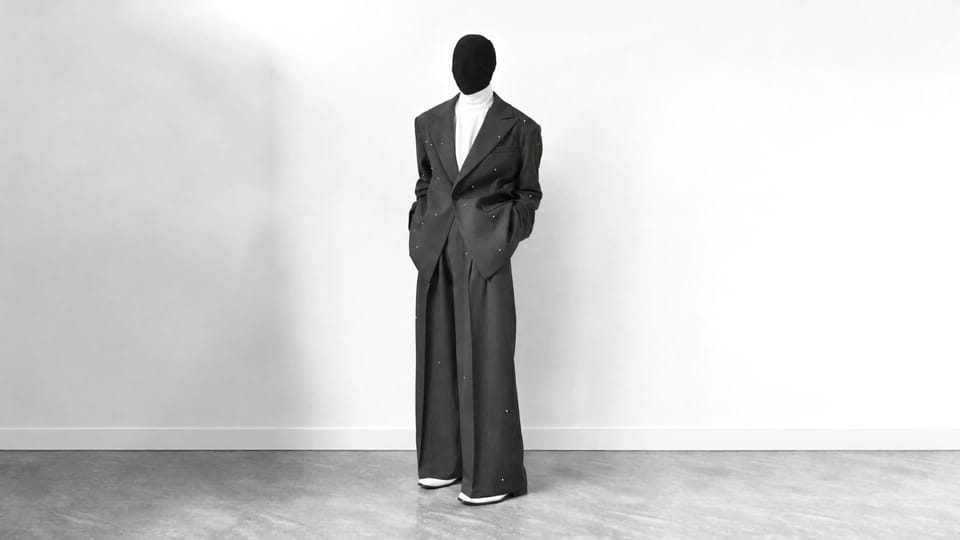 A standing figure in an oversized charcoal suit set with pearls, against a pale studio wall